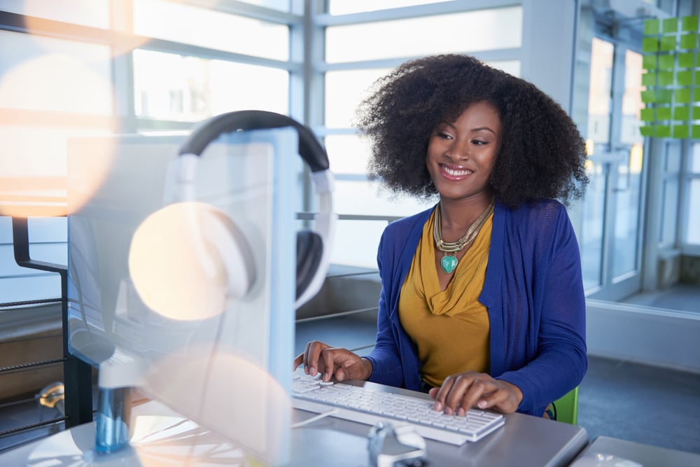Portrait of a smiling Black woman with an afro at the computer in bright glass office